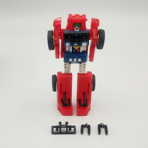 May include: A red and blue toy robot with black wheels and silver legs. The robot has a blue chest with a yellow triangle and a green face. Three small black accessories are at the bottom.