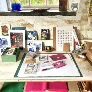 Op de afbeelding: Een werkruimte met een witte tafel bedekt met knutselbenodigdheden, waaronder kaarten, enveloppen en papier. Er zijn stapels kaarten met verschillende ontwerpen, waaronder vlinders, bloemen en dieren. Er zijn ook rollen tape, een marker en een roze envelop. De tafel is klaar voor verpakking en verzending.