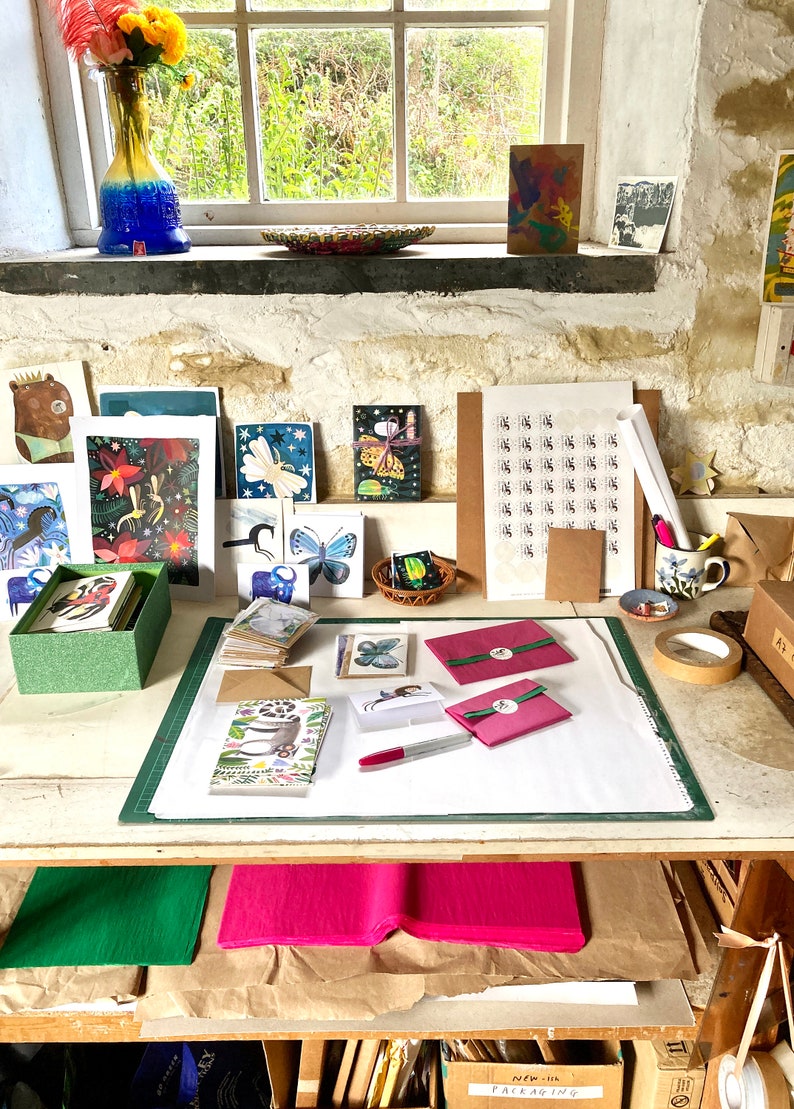 Op de afbeelding: Een werkruimte met een witte tafel bedekt met knutselbenodigdheden, waaronder kaarten, enveloppen en papier. Er zijn stapels kaarten met verschillende ontwerpen, waaronder vlinders, bloemen en dieren. Er zijn ook rollen tape, een marker en een roze envelop. De tafel is klaar voor verpakking en verzending.