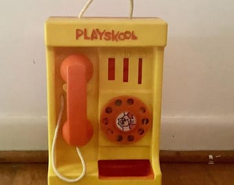 Könnte beinhalten: Ein gelbes Playskool-Spielzeugtelefon mit orangefarbenem Hörer und Wählscheibe. Das Telefon hat ein weißes Kabel und einen Griff oben. Das Wort "Playskool" ist oben aufgedruckt.