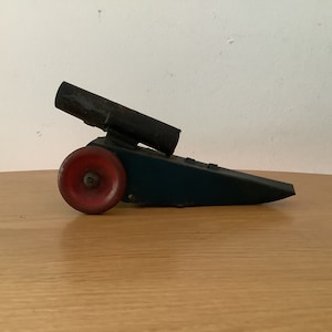 Może przedstawiać: Antyczne, zabawkowe działo z czarną metalową lufą i czerwonym drewnianym kołem. Działo jest zamontowane na niebieskiej drewnianej podstawie.