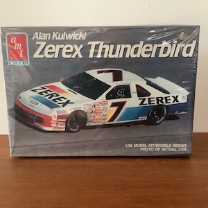 Puede incluir: Caja de modelo a escala del coche de carreras Alan Kulwicki Zerex Thunderbird. La caja presenta una imagen del coche, que es blanco con detalles en azul y rojo, el número 7 y logotipos de patrocinadores. El texto "Zerex Thunderbird" se muestra de forma destacada.