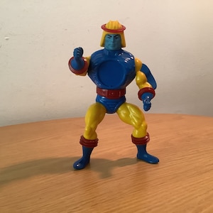 Puede incluir: Una figura de acción azul y amarilla con un casco rojo y amarillo. La figura está de pie sobre una superficie de madera.