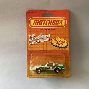 Puede incluir: Un coche Matchbox de fundición a presión, un Nissan 300ZX Turbo blanco y verde. El embalaje naranja y amarillo presenta el logotipo de Matchbox y el texto « The Automotive Superstars ». El juguete tiene piezas móviles.