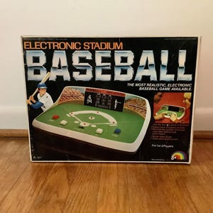 Może przedstawiać: Prostokątne kartonowe pudełko do gry Electronic Stadium Baseball. Na froncie znajduje się tytuł dużymi niebiesko-białymi literami, ilustracja baseballisty oraz grafika przedstawiająca zieloną elektroniczną konsolę do gier.