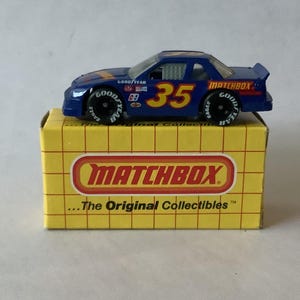 Puede incluir: Un coche de carreras Matchbox azul con el número 35 en amarillo, sobre una caja Matchbox amarilla. El coche tiene neumáticos Goodyear y logotipos de patrocinadores. La caja dice "MATCHBOX" y "The Original Collectibles."