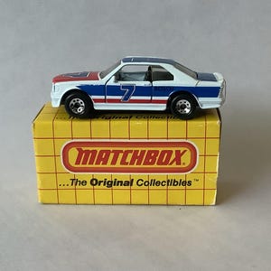 May include: A Matchbox die-cast model car, white with red and blue racing stripes, and the number 7. The car sits on a yellow Matchbox box with the text "MATCHBOX" and "...The Original Collectibles".