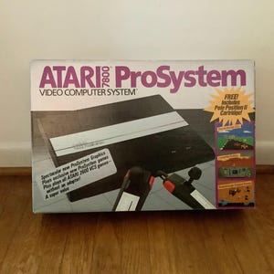 Könnte beinhalten: Die Verpackung des Atari 7800 ProSystem Video Computer Systems zeigt eine schwarze Konsole und zwei Joysticks mit roten Tasten. Die Schachtel trägt den Text Atari 7800 ProSystem, Video Computer System und enthält eine sternförmige Grafik mit der Aufschrift Free! Includes Pole Position II Cartridge!