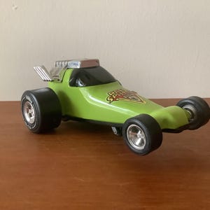 Op de afbeelding: Een groene speelgoed raceauto met zwarte wielen en een zilveren uitlaatpijp. De auto heeft de tekst "Baja Splinter" op de zijkant.