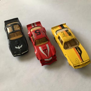 May include: Three vintage toy cars in black, red, and yellow. The black car has a bird emblem, the red car has the number 5, and the yellow car has the number 4 and the word "TURBO".
