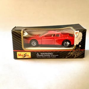 Könnte beinhalten: Rotes Modellauto Ferrari Testarossa in Originalverpackung. Die Verpackung ist schwarz mit goldenen Akzenten und enthält einen Warnhinweis auf Kleinteile. Sonderausgabe von Maisto.