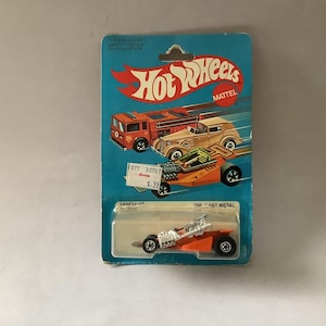 May include: A vintage Hot Wheels die-cast toy car on a blue and white card. The card features illustrations of orange and tan cars. The toy car is orange and silver, with black wheels. The card has the Hot Wheels logo.