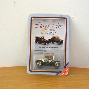 Peut inclure: Une voiture jouet verte avec un châssis noir et des accents marrons. La voiture est sur une carte en plastique avec le texte "Classic Cars Metal Collection Collect Them All Freewheel 301-306 Non-Toxic Paints Not Recommended for Children Under 3" imprimé dessus.