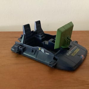 May include: A dark blue plastic G.I. Joe toy vehicle cockpit base featuring two molded seats, a hinged olive green radar screen, and a small American flag decal on the front hood. The base includes detailed paneling and a G.I. Joe logo sticker.