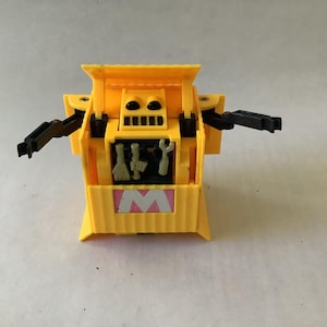 May include: A yellow plastic toy robot with black articulated arms and a rectangular body. The front features a central compartment containing small grey tools and a pink square panel with a white letter M printed in the center of the base.