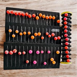 May include: A collection of fishing bait, featuring various colors and sizes of round, bead-like lures. The bait is arranged on black foam holders and a spool, with the brand name "FISHENG" visible. The lures are in shades of red, orange, and pink.