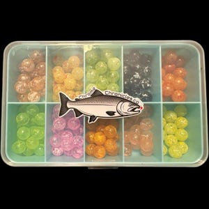 May include: A clear plastic tackle box with multiple compartments filled with colorful fishing beads. The beads are various colors, including orange, yellow, green, pink, and black. A sticker with a fish graphic and the text "Chrome Chronicles.co" is on the box.