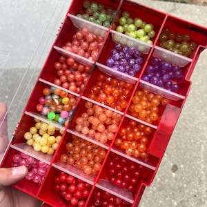 May include: A red plastic tackle box filled with small, colorful, round beads. The beads are arranged in rows and columns, and they are all different colors, including green, yellow, orange, red, and purple.