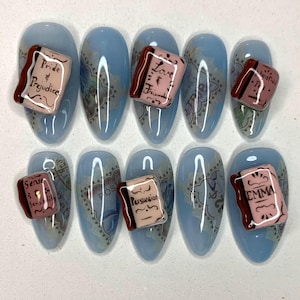 Puede incluir: Un conjunto de diez uñas almendradas de color azul claro. Cada uña presenta un dije de libro en miniatura con títulos como "Pride & Prejudice" y "Emma". Las uñas tienen un diseño de mapa vintage.