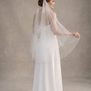 May include: A bride wearing a white wedding dress and a sheer veil. The veil is layered and falls over the shoulders and back. The dress is a simple, elegant design with a flowing skirt. The background is a neutral, light brown colour.