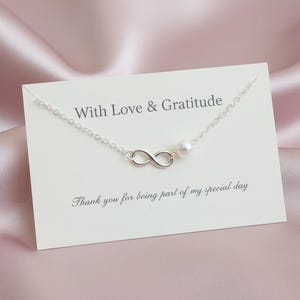 May include: A silver infinity necklace with a pearl pendant, presented on a white card. The card reads "With Love & Gratitude" and "Thank you for being part of my special day". The background is a pink satin material.