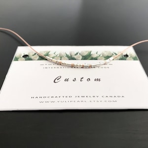 Peut inclure: Un collier personnalisé en gros plan avec des perles argentées et dorées sur un cordon rose. Le collier est présenté sur une carte blanche avec le texte "Custom" et "Handcrafted Jewelry Canada" imprimé dessus.