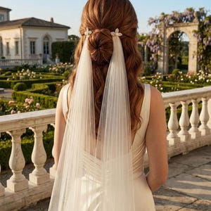 May include: A white bridal veil attached to two decorative hair clips featuring a central pearl surrounded by small clear crystals. The veil consists of two long, sheer panels of tulle fabric that drape down from a half-up hairstyle with a bun.