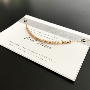 May include: A gold necklace with a delicate chain and small beads spelling out "Soul Sister" in Morse code. The necklace is displayed on a white card with the text "Handcrafted Jewelry Canada www.yulipearl.etsy.com".