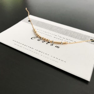 Peut inclure: Un collier en chaîne dorée avec un message personnalisé en perles dorées et argentées. Le collier est sur une carte blanche avec le texte "HANDCRAFTED JEWELRY CANADA WWW.YULINEAR.COM" et "INTERNATIONAL MORSE CODE" écrit dessus.