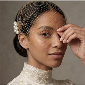 May include: A gold-toned birdcage veil featuring a fine mesh netting that covers the upper face. The veil is secured on one side with a cluster of six smooth, round, white faux pearls arranged in a vertical row against the hair.