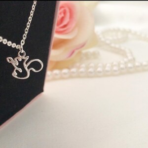 May include: A silver necklace featuring a squirrel pendant. The pendant is a minimalist outline. The necklace is presented against a black backdrop, with a pink rose and a string of pearls visible in the background.