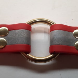 Puede incluir: Un collar rojo y plateado con un anillo de metal dorado en el centro. El collar tiene dos secciones con cierres de presi&oacute;n de metal dorado.