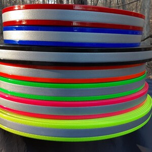 Puede incluir: Una pila de rollos de cincha reflectante de colores. Los rollos presentan una franja reflectante plateada con bordes de varios colores, como rojo, azul, negro, naranja, verde y rosa. La cincha se utiliza probablemente para fines de seguridad o visibilidad.