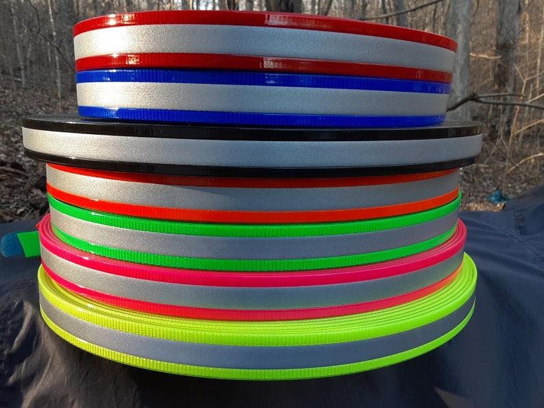 Puede incluir: Una pila de rollos de cincha reflectante de colores. Los rollos presentan una franja reflectante plateada con bordes de varios colores, como rojo, azul, negro, naranja, verde y rosa. La cincha se utiliza probablemente para fines de seguridad o visibilidad.