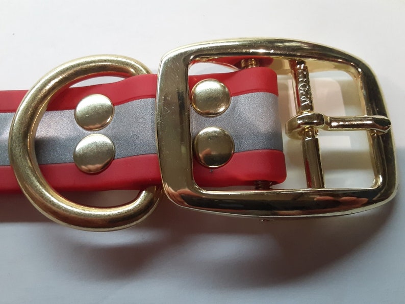 Puede incluir: Primer plano de un collar para perros con un dise&ntilde;o a rayas rojas y plateadas. El collar presenta herrajes met&aacute;licos dorados, que incluyen una hebilla, una anilla en D y remaches. El collar est&aacute; hecho de un material flexible.