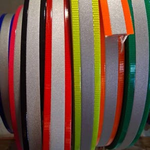 Puede incluir: Primer plano de varios rollos de cinta reflectante en varios colores. Los rollos incluyen plata, negro, rojo, naranja, verde y amarillo. La cinta se utiliza probablemente con fines de seguridad o decorativos.