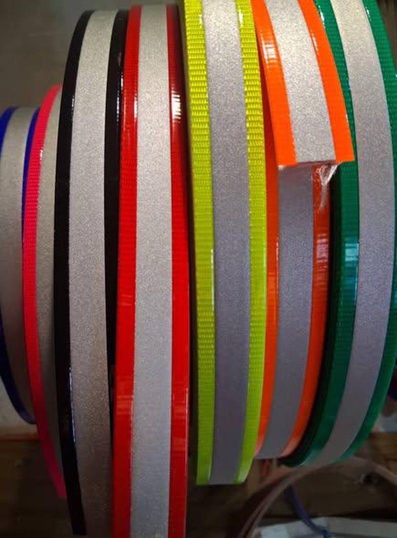 Puede incluir: Primer plano de varios rollos de cinta reflectante en varios colores. Los rollos incluyen plata, negro, rojo, naranja, verde y amarillo. La cinta se utiliza probablemente con fines de seguridad o decorativos.