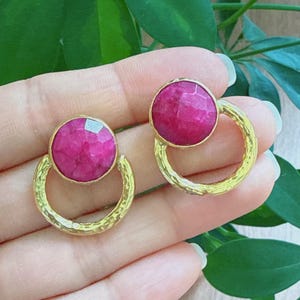 Puede incluir: Un par de pendientes dorados con una gema magenta redonda y facetada, colocada sobre un diseño de semicírculo texturizado. Los pendientes se muestran sobre un fondo de follaje verde.