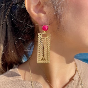 May include: Gold earrings with a geometric design and a pink stone accent.