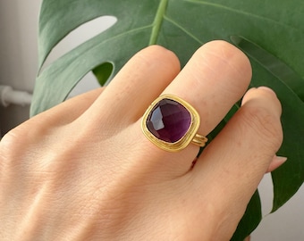 May include: A gold ring with a square, deep purple gemstone. The ring has a thick gold band and a gold bezel setting around the gemstone. The ring is on a person's finger, with a green leaf in the background.