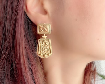 May include: Gold-colored, ornate earrings with a rectangular top and a decorative, elongated bottom. The earrings feature detailed floral and geometric patterns. The image shows a close-up of the earrings on a person's ear, highlighting the intricate design and warm metallic hue.