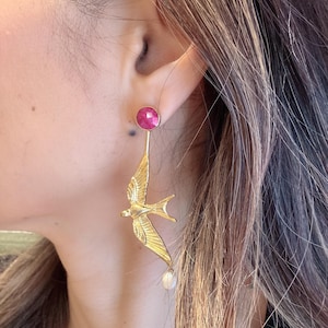 Può includere: Orecchini a forma di uccello color oro con borchia in pietra rosa e perla pendente. Gli orecchini sono indossati sull'orecchio di una persona, mettendo in risalto gli intricati dettagli del design dell'uccello e il contrasto tra oro, rosa e bianco.