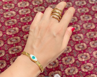 May include: A gold bracelet with a white and blue evil eye charm. The bracelet is on a person's wrist. The person is wearing a gold ring on their finger.