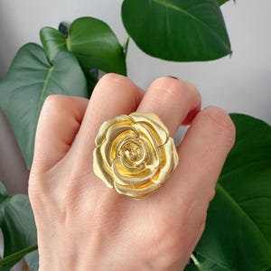 May include: A gold-colored ring shaped like a rose. The ring is large and has a detailed design.