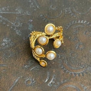 May include: A gold-toned ring featuring a branch-like design adorned with several faux pearls. The ring has a textured, organic appearance, with the pearls set at intervals along the branches. The ring is set against a dark, textured background.