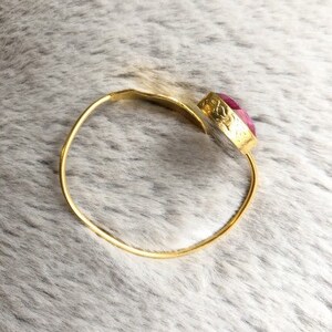 May include: A gold-colored bangle bracelet with a large, oval, pink gemstone. The gemstone is set in a decorative gold bezel. The bracelet is simple and elegant, suitable for everyday wear or special occasions.