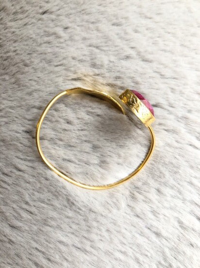 May include: A gold-colored bangle bracelet with a large, oval, pink gemstone. The gemstone is set in a decorative gold bezel. The bracelet is simple and elegant, suitable for everyday wear or special occasions.