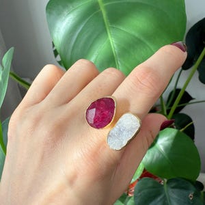 Puede incluir: Un anillo de oro con dos piedras. La primera piedra es de color rojo intenso, y la segunda piedra es un cuarzo blanco y nublado.
