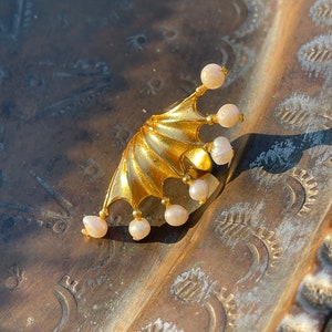 May include: A gold ear cuff in a bat wing shape, embellished with small, round pearls. The cuff is set against a textured, bronze-coloured surface, creating a striking contrast. This unique piece of jewellery adds a touch of elegance.