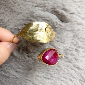 May include: Two gold rings. One ring is a leaf shape with a small, round, clear stone. The other ring has a teardrop-shaped, deep red stone. Both rings have a gold band. The rings are displayed on a gray, textured surface.